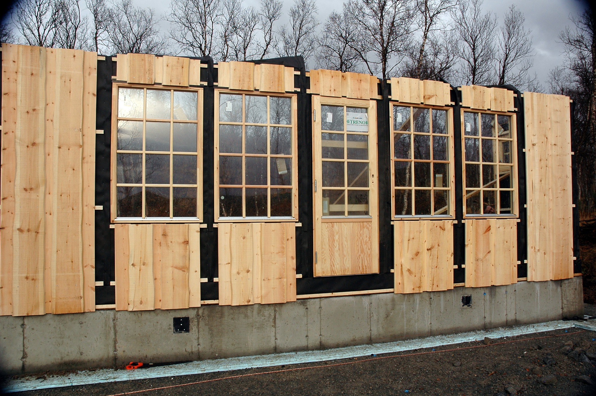 Montaż drewnianej elewacji z oknami i drzwiami, widoczne deski z surowego drewna na czarnej membranie, betonowa podmurówka, drzewa w tle.