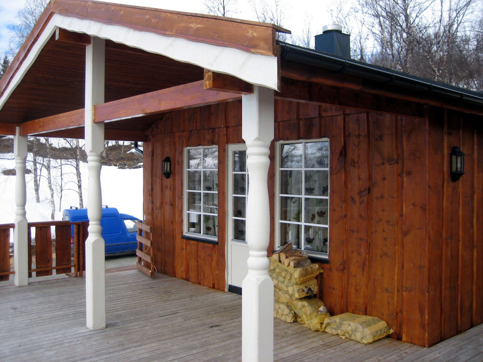 Drewniany dom z werandą, widoczne białe kolumny, okna z białymi ramami, sterta drewna przy ścianie, na zewnątrz stoi niebieski samochód dostawczy.