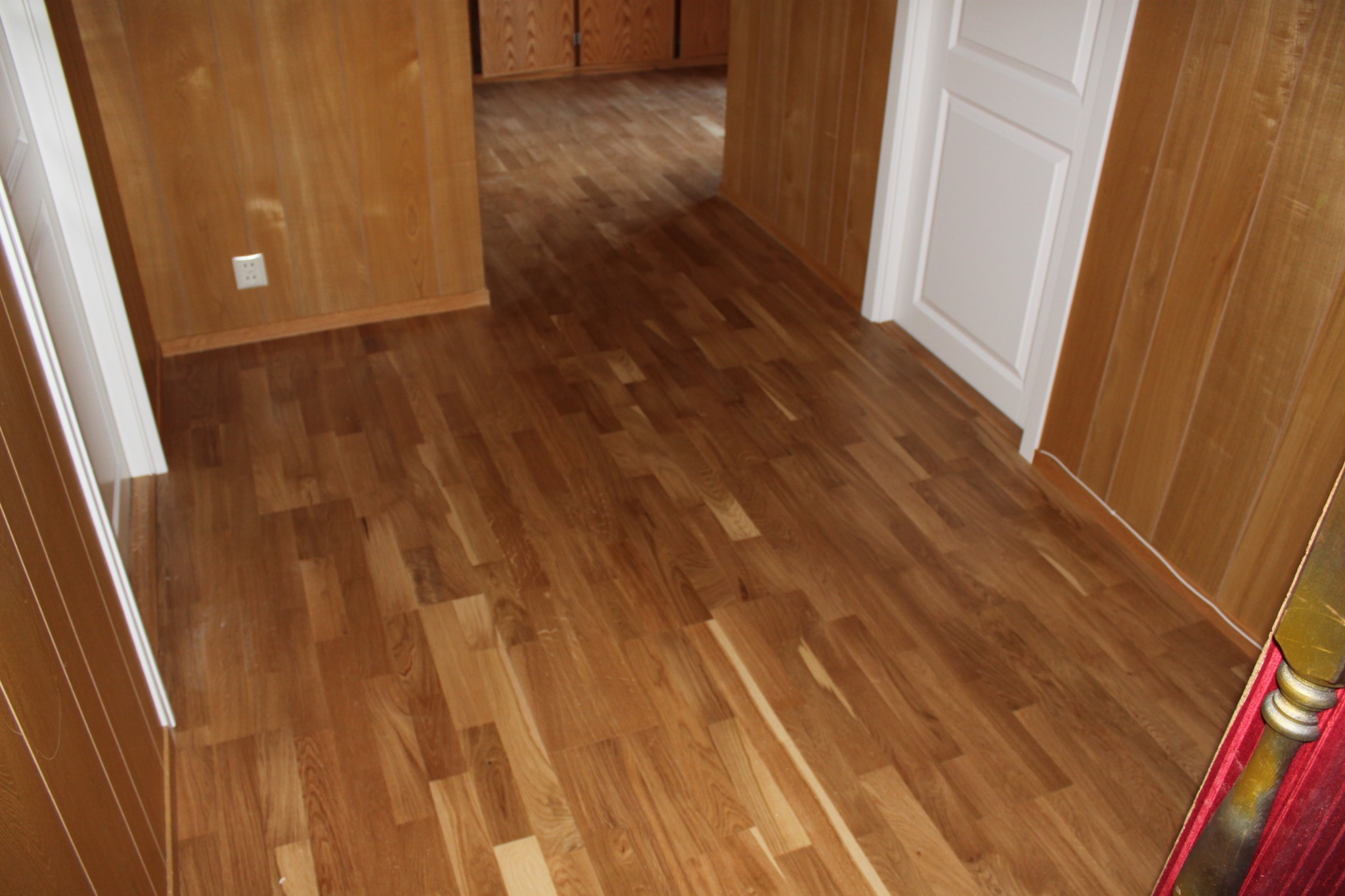 Wnętrze z panelami ściennymi z drewna i podłogą z paneli w odcieniach brązu, widoczny biały kontakt i fragment białych drzwi.