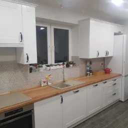 Jasna kuchnia z białymi frontami szafek i drewnianym blatem, widok na okno z dekoracjami świątecznymi, szare płytki na ścianie i podłodze.