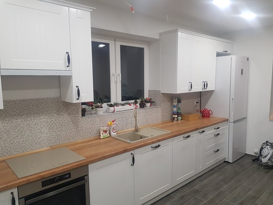 Jasna kuchnia z białymi frontami szafek i drewnianym blatem, widok na okno z dekoracjami świątecznymi, szare płytki na ścianie i podłodze.