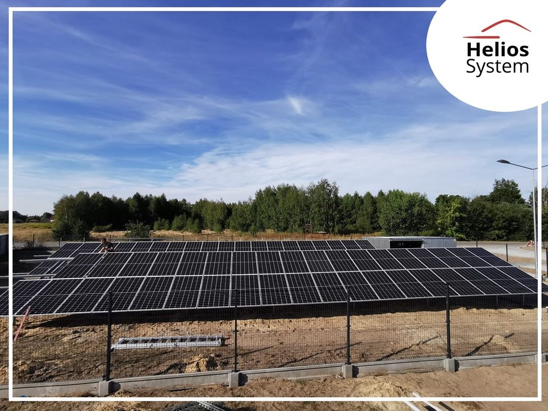Instalacja fotowoltaiczna na ziemi, widok zza ogrodzenia, błękitne niebo z delikatnymi chmurami, w tle drzewa i słup oświetleniowy, w prawym górnym rogu logo firmy Helios System.
