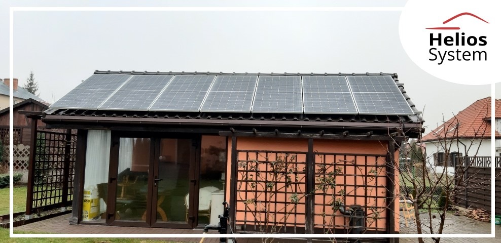 Panele słoneczne na dachu małego budynku w ogrodzie, widoczne logo firmy Helios System w prawym górnym rogu.