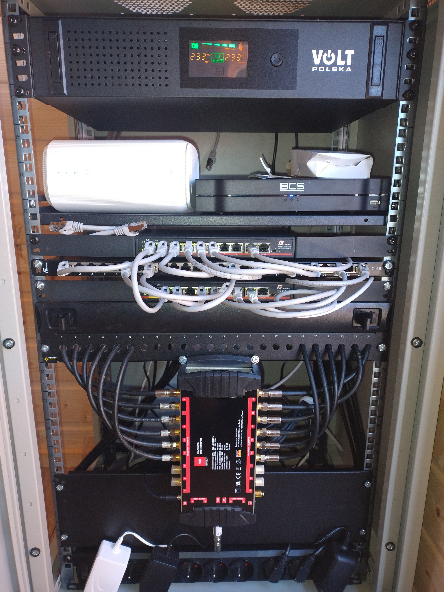 Szafa rack z zainstalowanymi urządzeniami: UPS Volt Polska, router WiFi, przełączniki sieciowe z podłączonymi kablami Ethernet, multiswitch z kablami koncentrycznymi, listwa zasilająca...