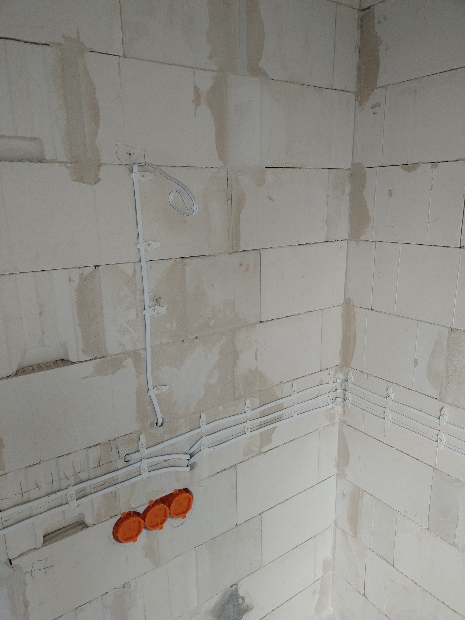 Surowe ściany z betonu komórkowego z zamontowanymi białymi rurkami instalacji elektrycznej i trzema pomarańczowymi puszkami pod gniazdka.