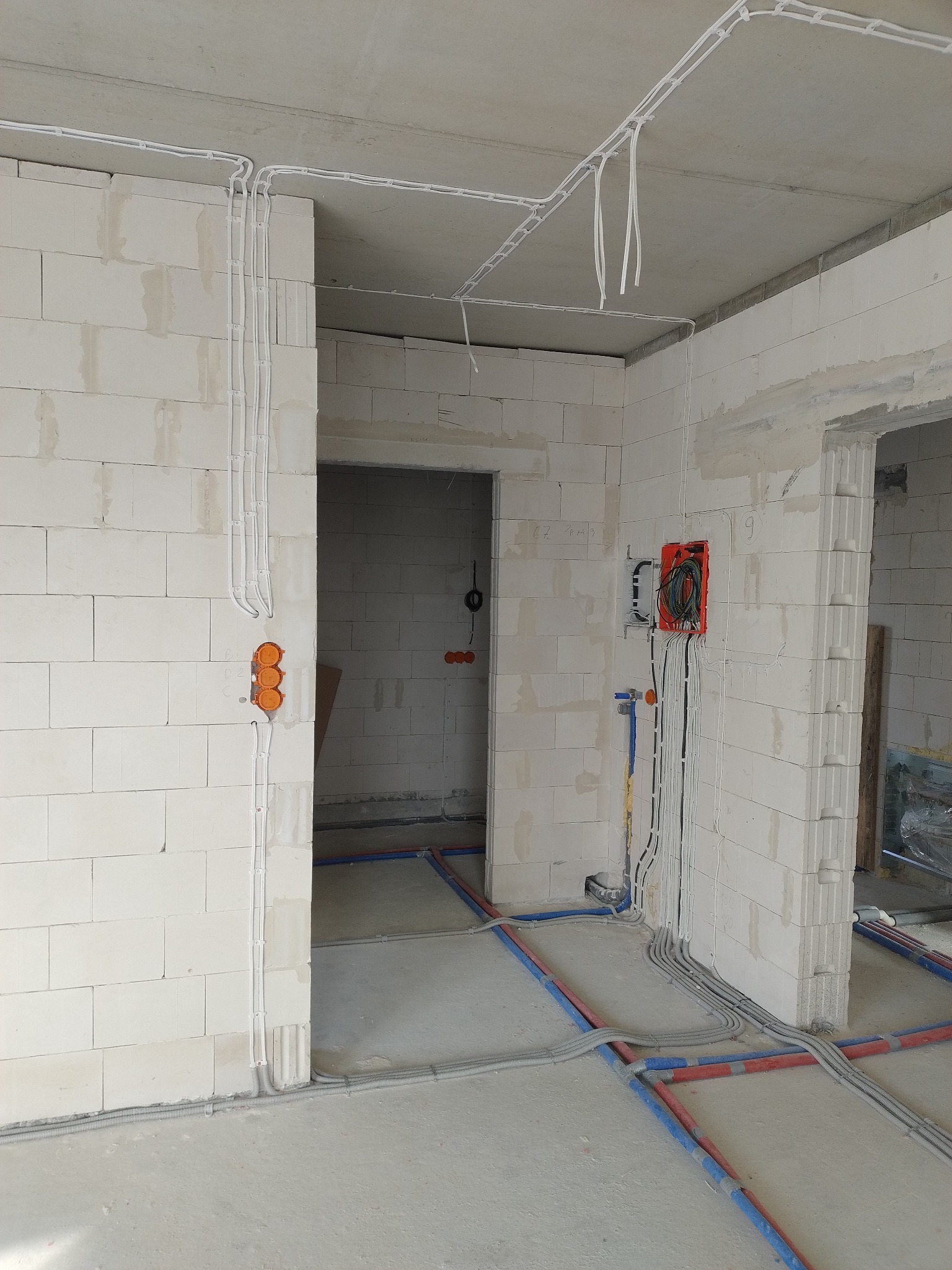 Surowy stan wnętrza z widocznymi przewodami elektrycznymi i rurami rozprowadzonymi po ścianach i podłodze, puszki instalacyjne w ścianach z betonu komórkowego.