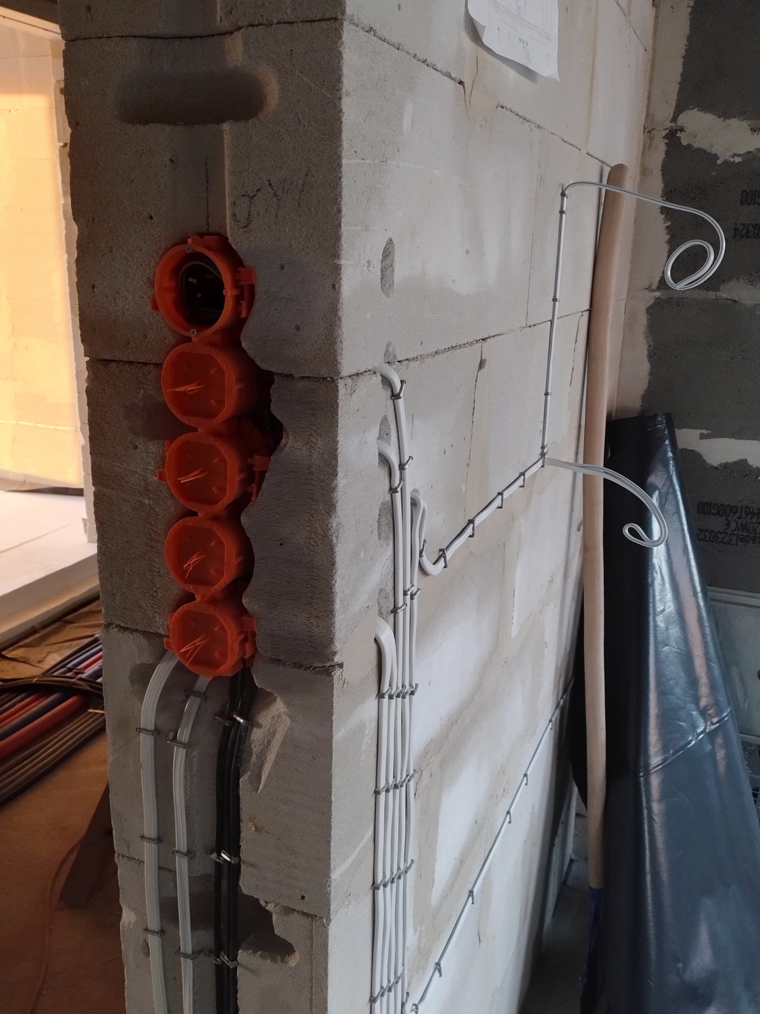 Surowy mur z betonu komórkowego z zamontowanymi, pomarańczowymi puszkami elektrycznymi pod gniazdka i wciągniętymi przewodami. Kable prowadzone wzdłuż ściany, mocowane metalowymi uchwytami.
