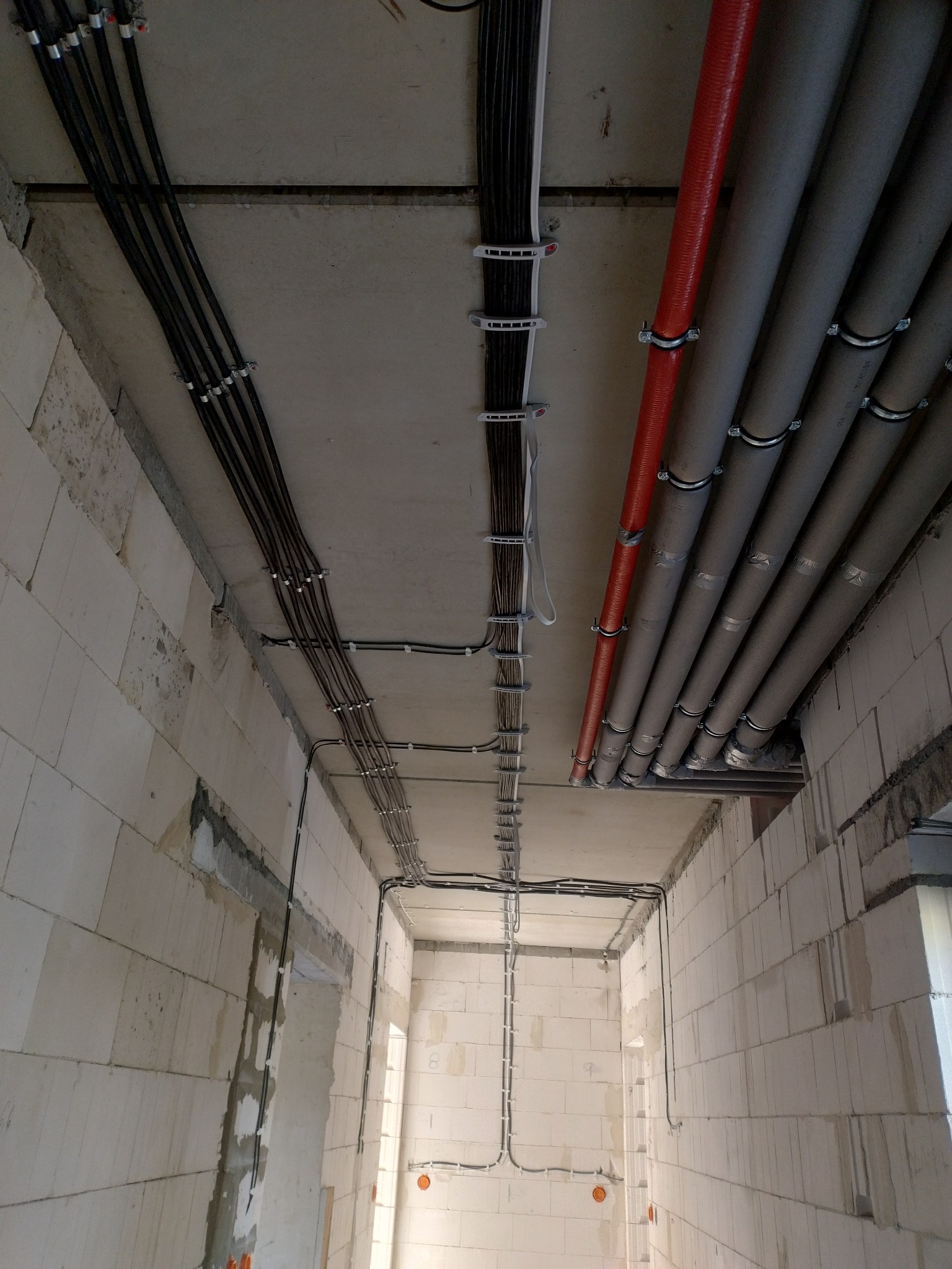 Wnętrze w stanie surowym z widocznymi przewodami elektrycznymi i rurami wentylacyjnymi, zamocowanymi do sufitu i ścian za pomocą uchwytów.