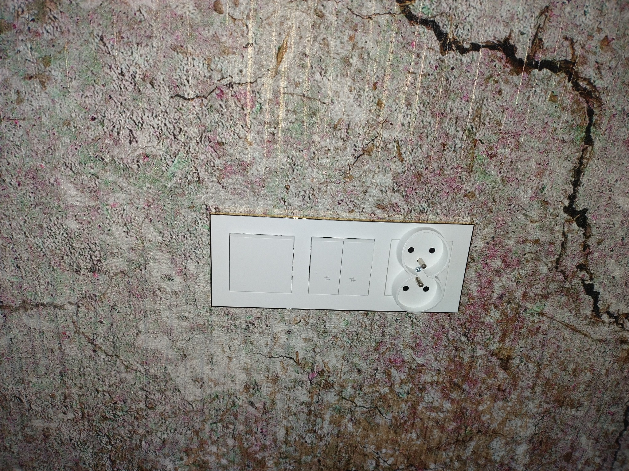 Biały panel z dwoma włącznikami i podwójnym gniazdkiem elektrycznym zamontowany na ścianie o nieregularnej, wzorzystej fakturze przypominającej korę drzewa.