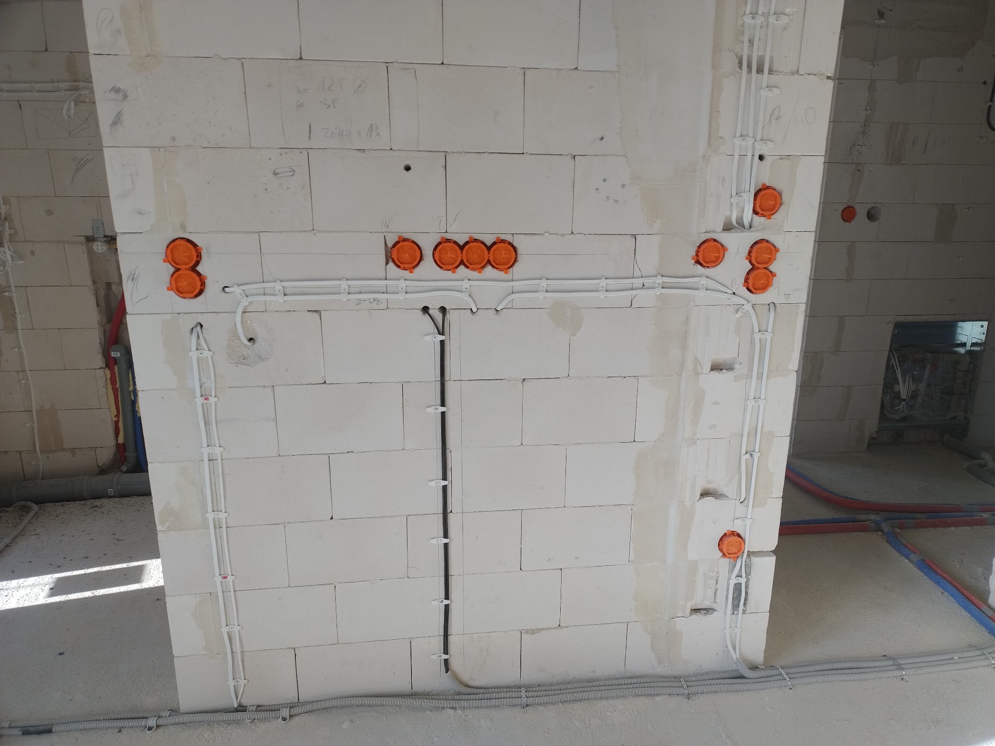 Surowa ściana z betonu komórkowego z widocznymi puszkami elektrycznymi w kolorze pomarańczowym i przewodami w białych peszlach, instalacja w trakcie montażu.