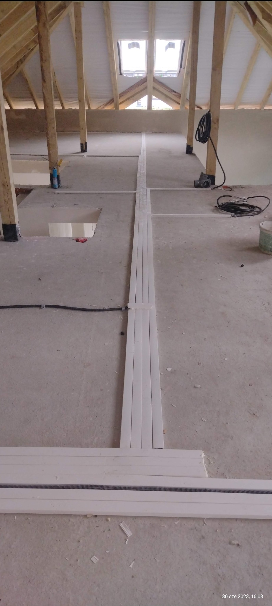 Widok poddasza w trakcie prac, z widocznymi elementami instalacji elektrycznej w postaci białych listew przypodłogowych z kanałami kablowymi oraz przewodami elektrycznymi prowadzonymi po podłodze.