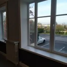 Białe okno PCV z widokiem na parking i samochód, ściana pomalowana na ciemny brąz, widoczny fragment grzejnika i parapetu.