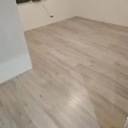 Jasne panele podłogowe ułożone we wzór imitujący drewno w pustym pokoju z beżowymi ścianami. Widoczny fragment białej ściany z lewej strony i gniazdko elektryczne na ścianie. Na podłodze widoczne...