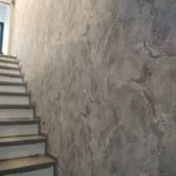 Ściana wewnętrzna z dekoracyjną fakturą imitującą kamień, przylegająca do schodów z szarymi stopniami i białymi podstopnicami. Widoczne gniazdka elektryczne na dole ściany.