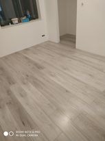 Jasnoszare panele podłogowe ułożone w pustym, jasnym pomieszczeniu. Widoczny fragment okna z parapetem, na którym stoją pojemniki. Świeżo położona podłoga.