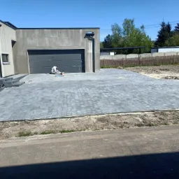 Ułożona szara kostka brukowa przed nowoczesnym domem z garażem; pracownik na kolanach, układający ostatnie elementy nawierzchni, widoczne narzędzia i piasek do fugowania.