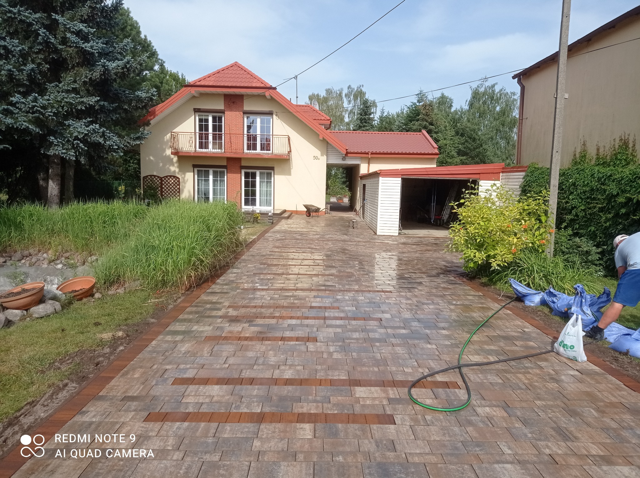 Nowo ułożony, wzorzysty podjazd z kostki brukowej prowadzący do domu z czerwoną dachówką, widoczny wąż ogrodowy i częściowo zakryte worki z materiałem.
