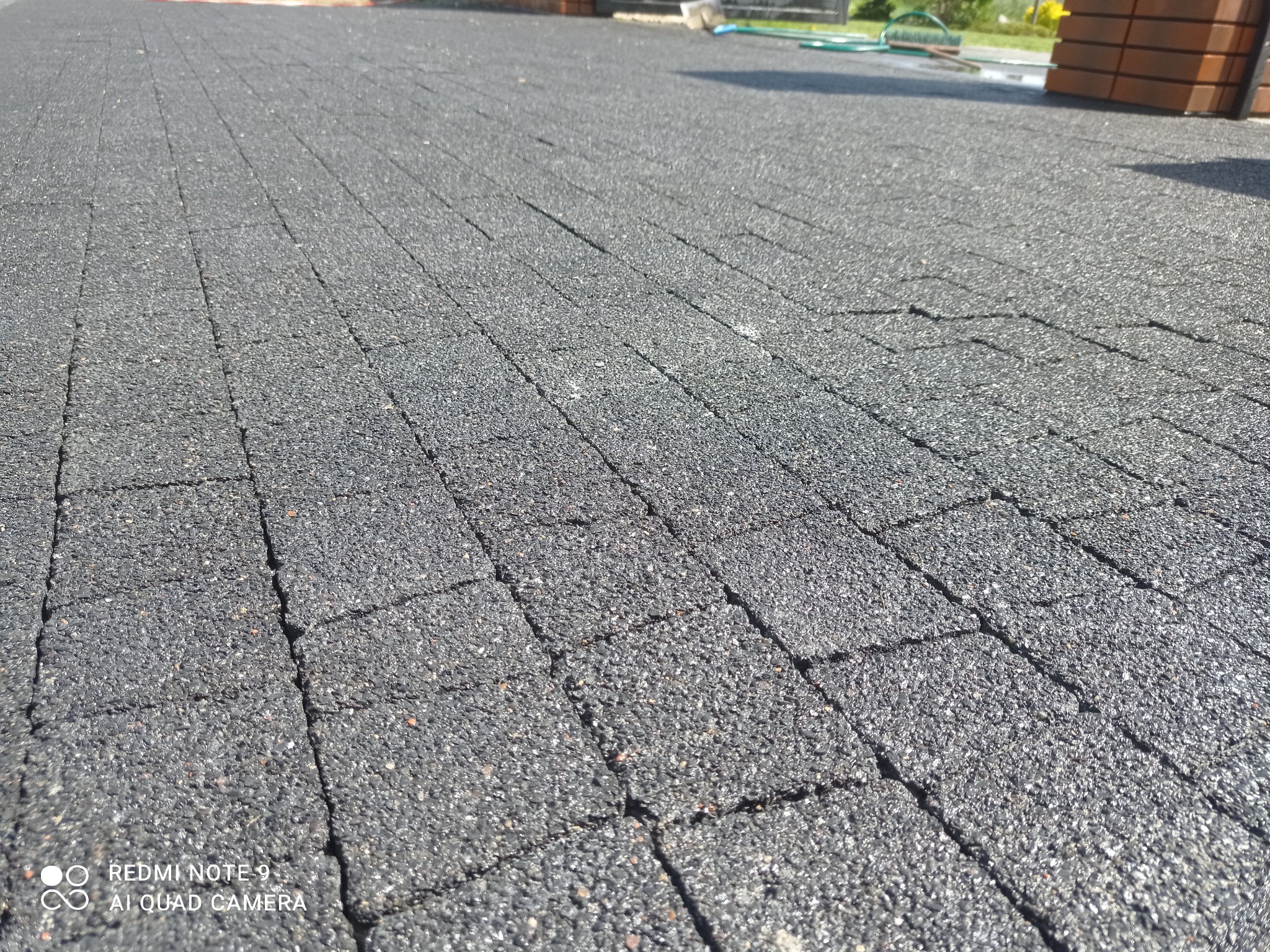 Szara, porowata kostka brukowa ułożona na posesji, widoczna z niskiej perspektywy, z elementami ogrodu w tle.