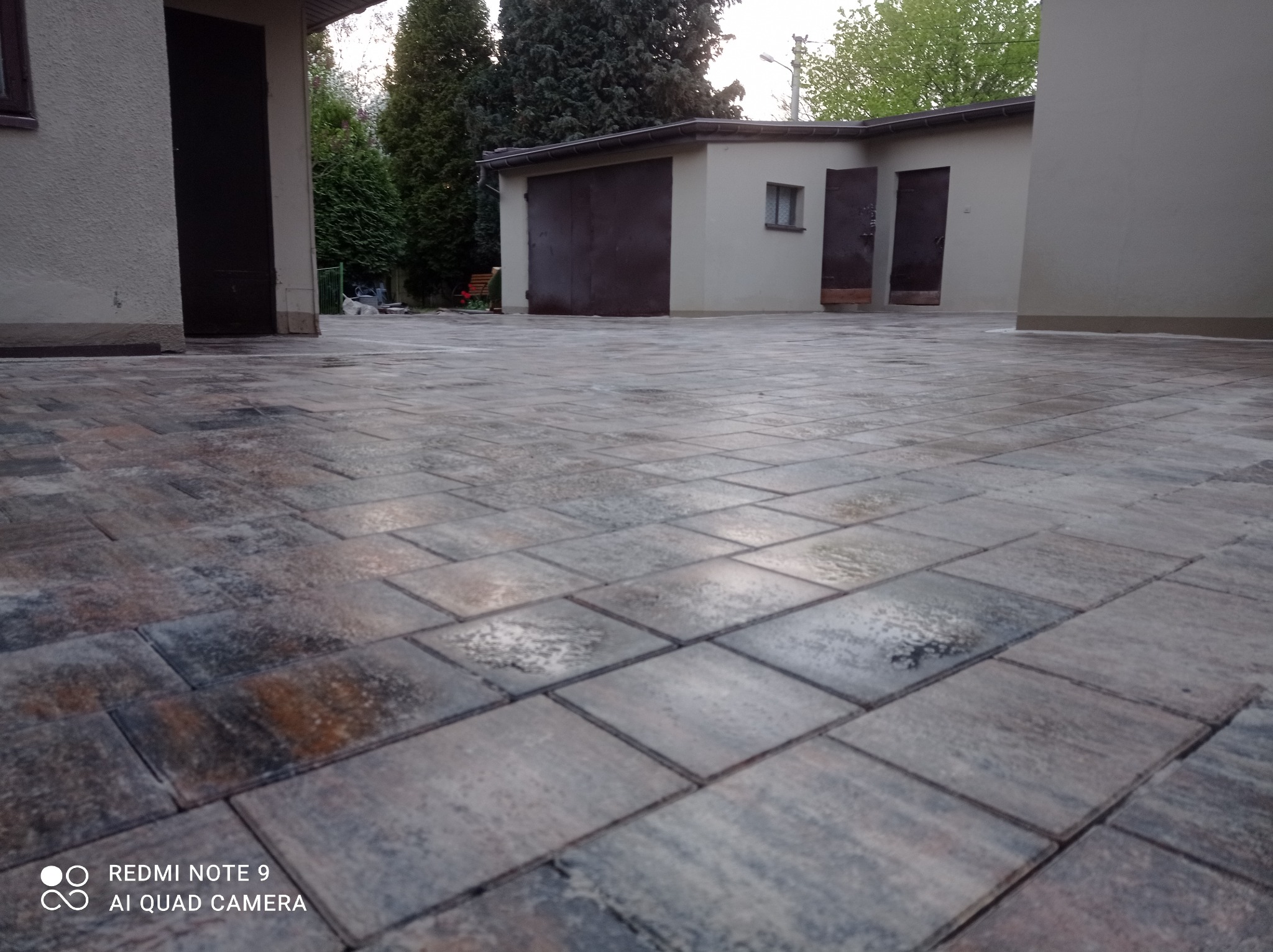 Szara kostka brukowa ułożona na podjeździe przed garażem i budynkiem gospodarczym w pochmurny dzień.