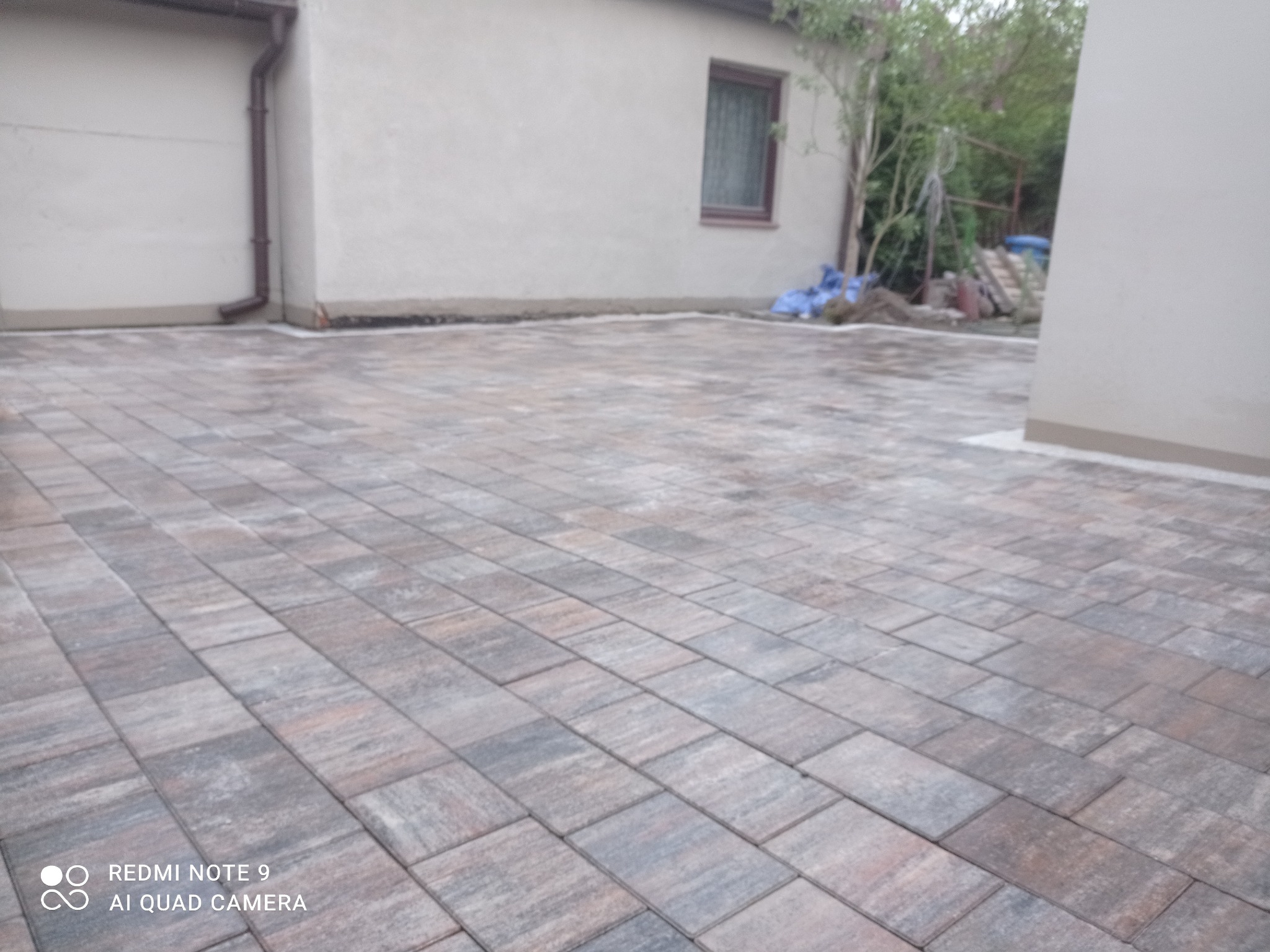 Ułożona kostka brukowa w odcieniach szarości i brązu przed budynkiem z widoczną rynną spustową i fragmentem ogrodu w tle.