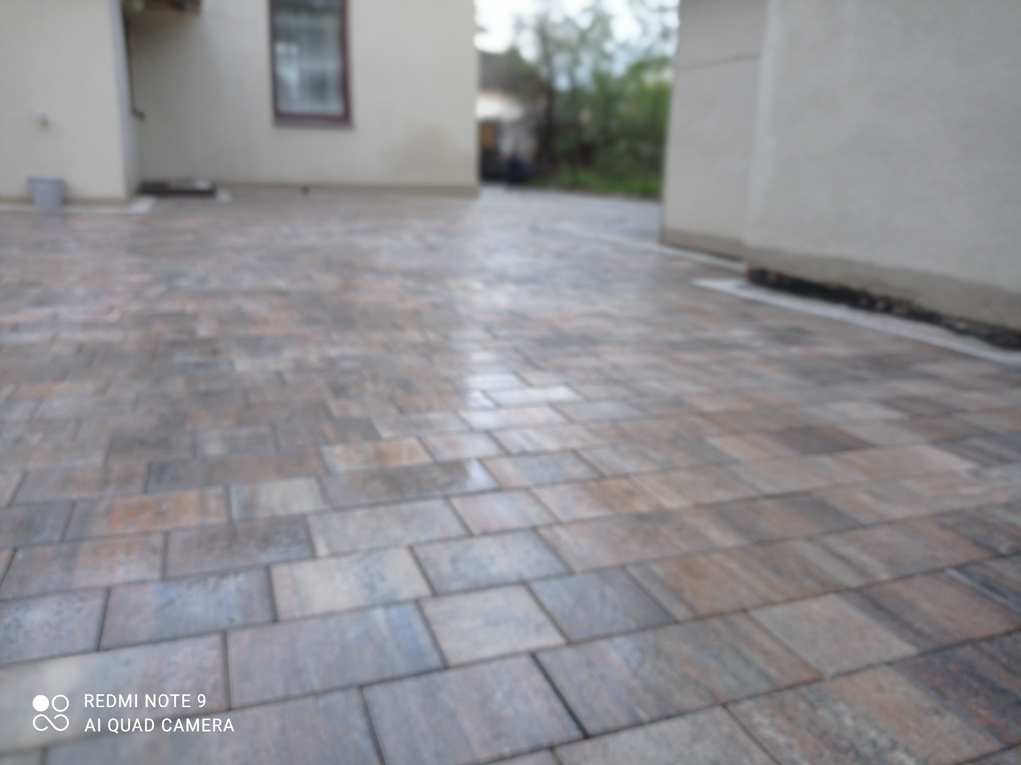 Świeżo ułożona, mokra kostka brukowa w odcieniach szarości i brązu, tworząca chodnik przed budynkiem o jasnej elewacji. Widoczne odbicia światła na powierzchni kostki.