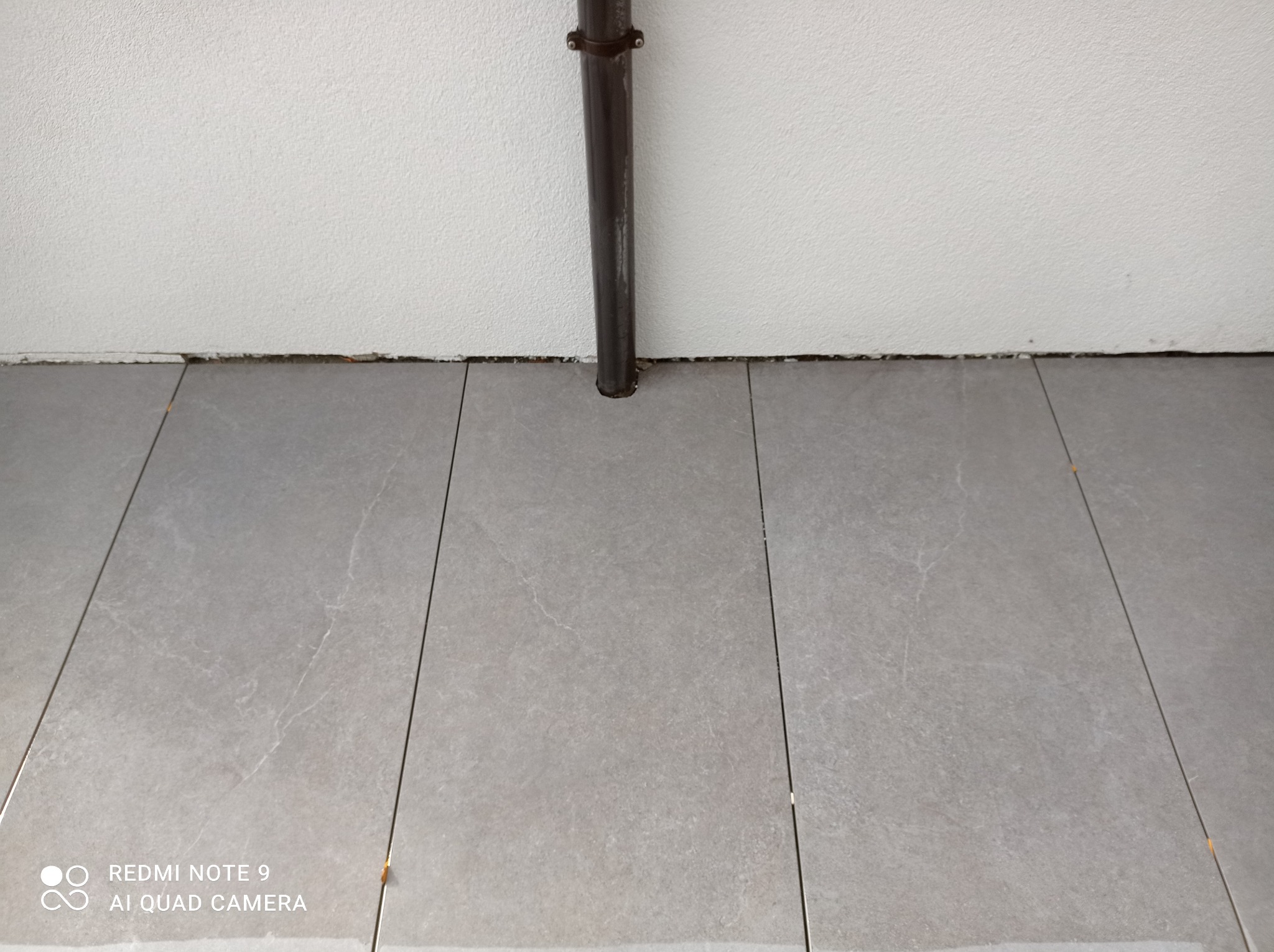 Szare płytki podłogowe ułożone wzdłuż ściany z widoczną rurą pionową, brak listew przypodłogowych.