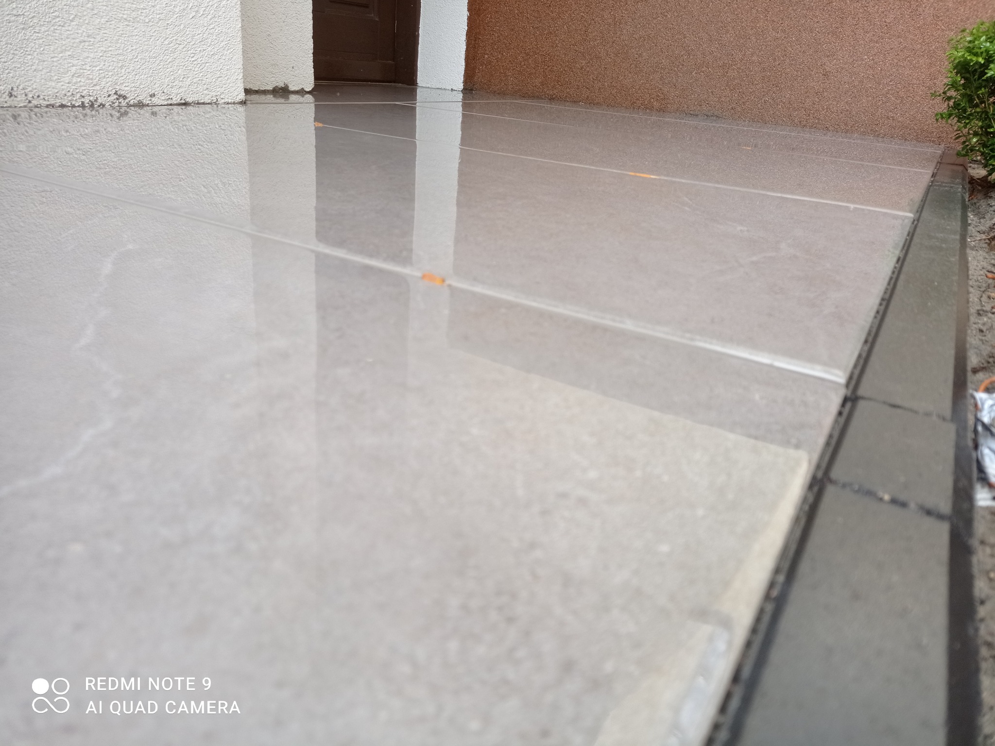 Świeżo ułożona, błyszcząca, jasnoszara kostka brukowa przed wejściem do budynku, odbijająca światło, z widocznymi fugami i fragmentem zieleni po prawej stronie.