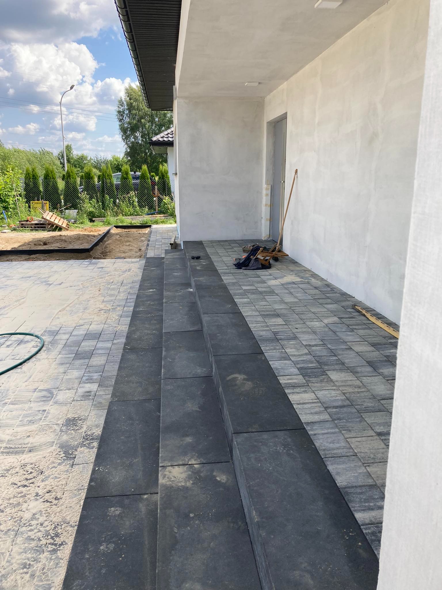 Ułożona kostka brukowa w odcieniach szarości prowadząca do wejścia domu, z widocznymi schodami z ciemnej kostki i przygotowanym podłożem pod dalszą część nawierzchni.