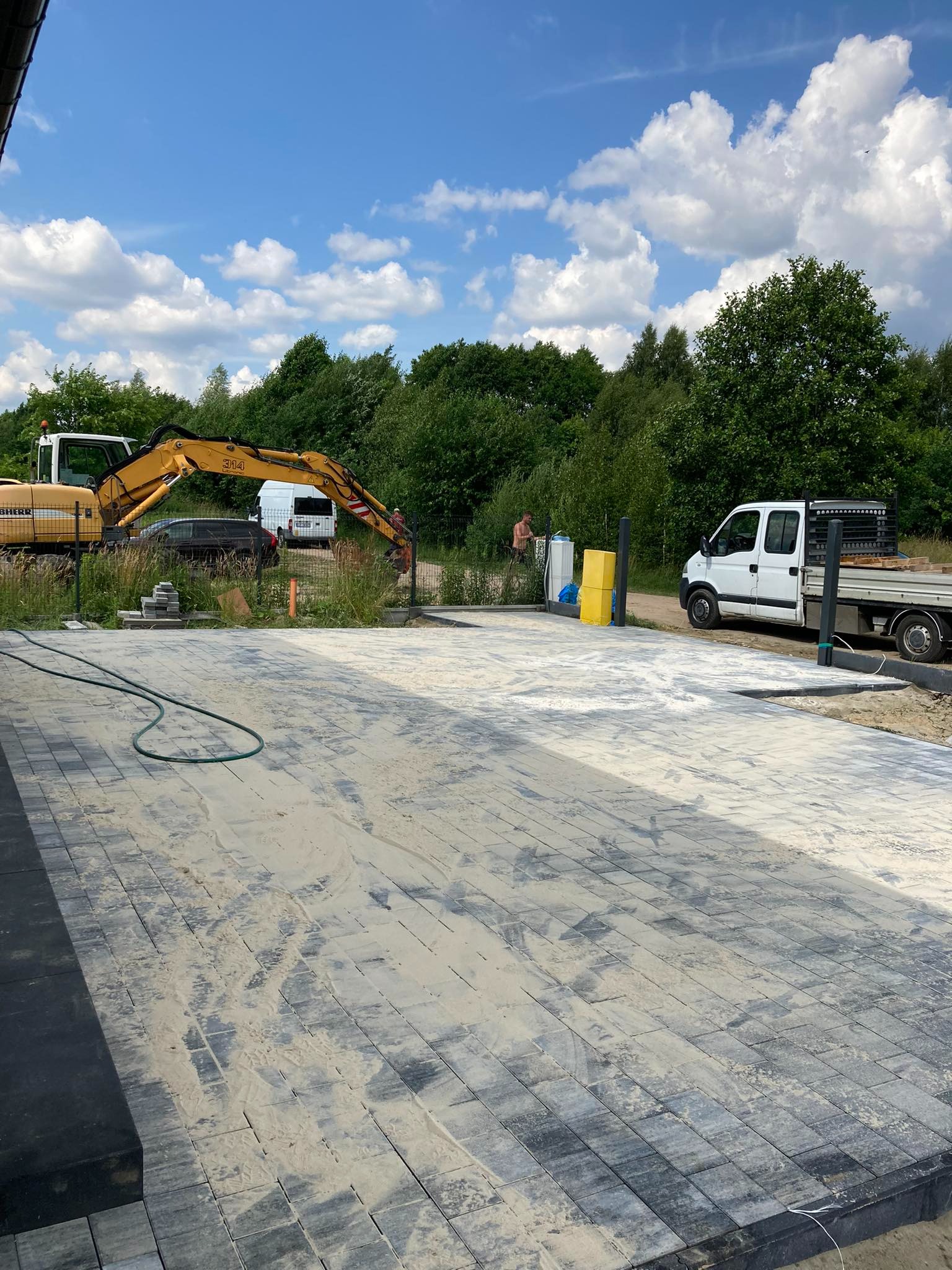 Nowo ułożona kostka brukowa w odcieniach szarości, pokryta piaskiem, z widocznym w tle żółtym sprzętem budowlanym i ciężarówką dostawczą, na tle zieleni i błękitnego nieba z chmurami.