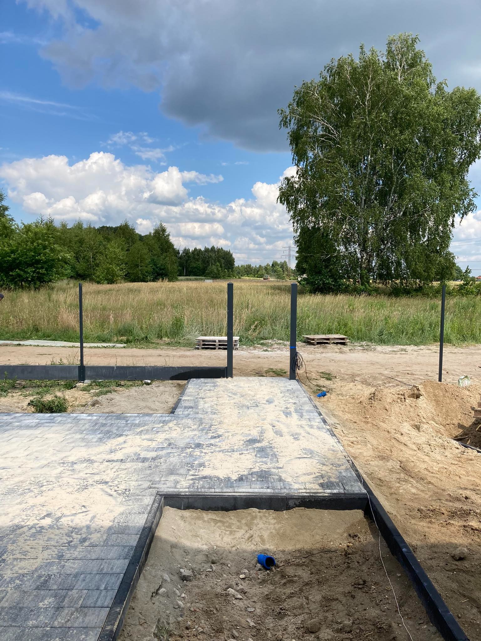 Fragment nowej nawierzchni z kostki brukowej, częściowo zasypanej piaskiem, z przygotowanym obramowaniem pod dalsze prace, słupki ogrodzeniowe w tle i widok na łąkę i drzewa w słoneczny dzień.