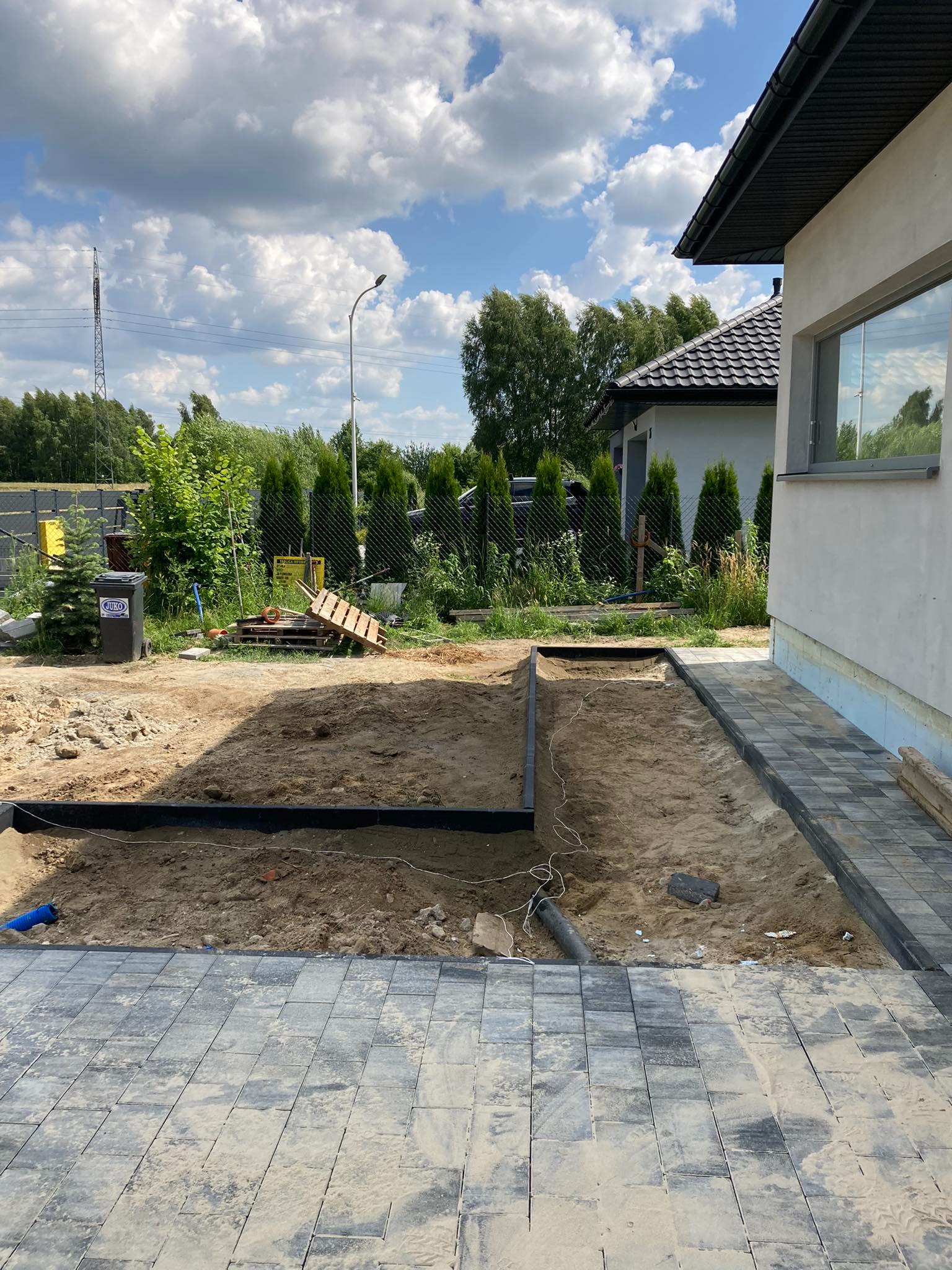 Przygotowanie podłoża pod kostkę brukową: wyrównany piasek z obrzeżami i fragmentem ułożonej nawierzchni przy ścianie budynku, widoczne narzędzia i materiały budowlane w tle.