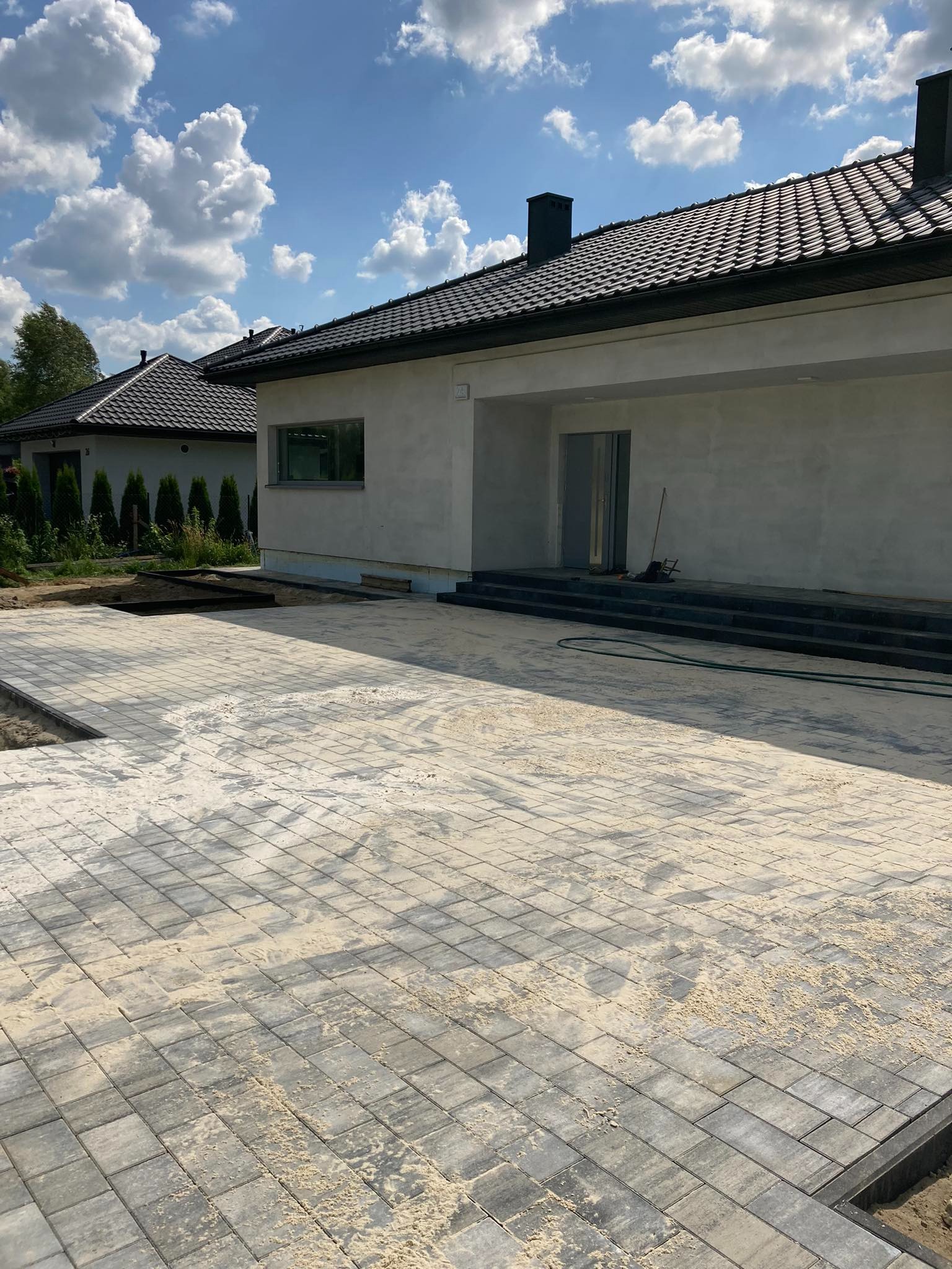 Ułożona kostka brukowa w odcieniach szarości, zasypana piaskiem na podjeździe przed nowoczesnym domem z ciemnym dachem i schodami wejściowymi.
