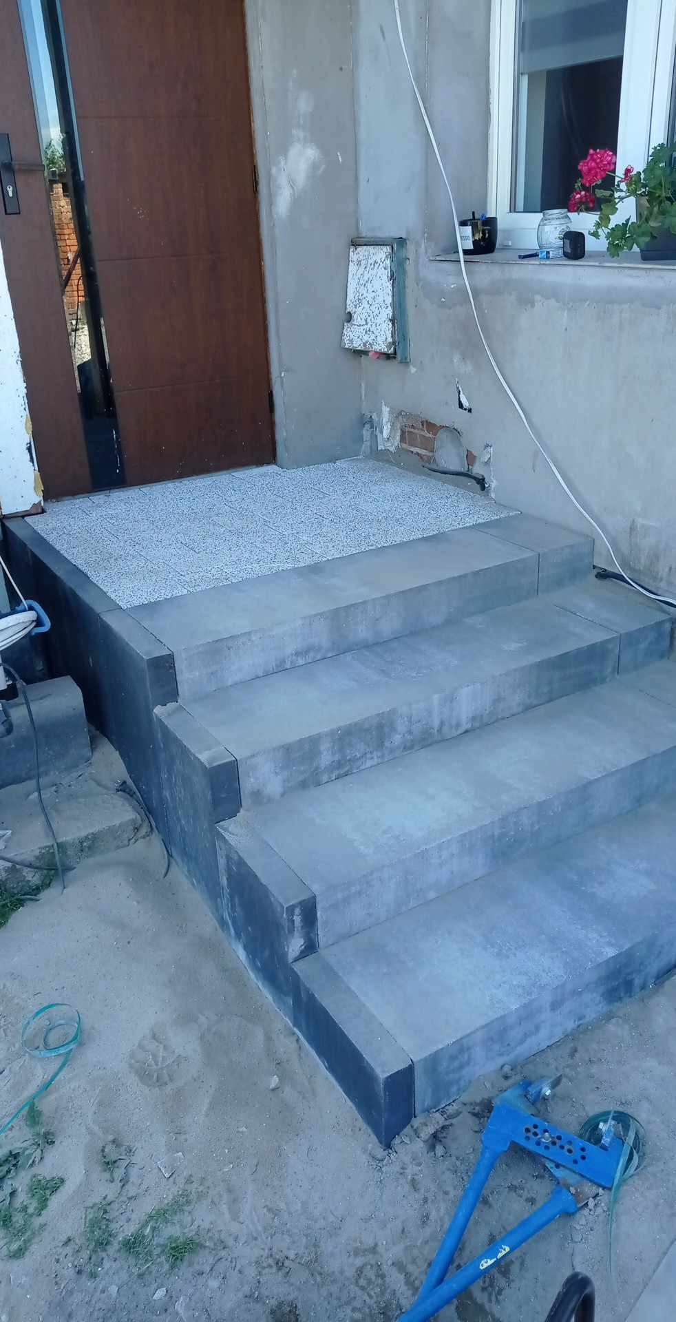 Nowe, betonowe schody wejściowe prowadzące do domu, zakończone płytkami na górnym podeście, z widocznym zagęszczarką w tle.