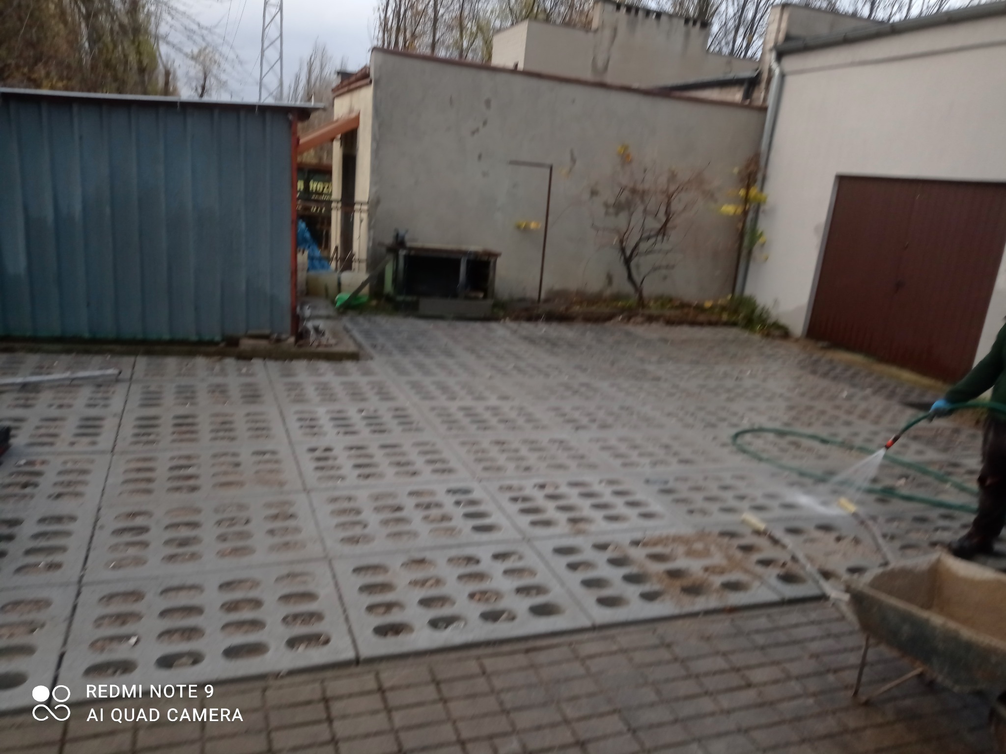 Szara, ażurowa kostka brukowa na podwórku, czyszczona wodą z węża, obok taczka, w tle budynki gospodarcze i drzewa bez liści.