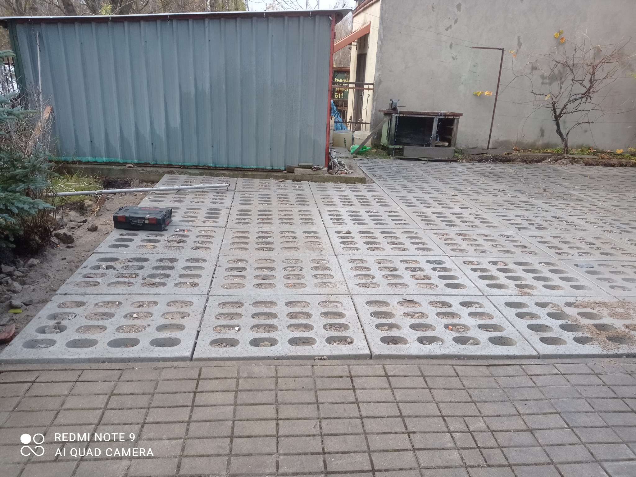 Ułożona nawierzchnia z ażurowych betonowych płyt na podwórku, częściowo wypełnionych ziemią, z widocznym narzędziem w czarnej skrzynce, metalowym garażem i fragmentem starego budynku w tle.