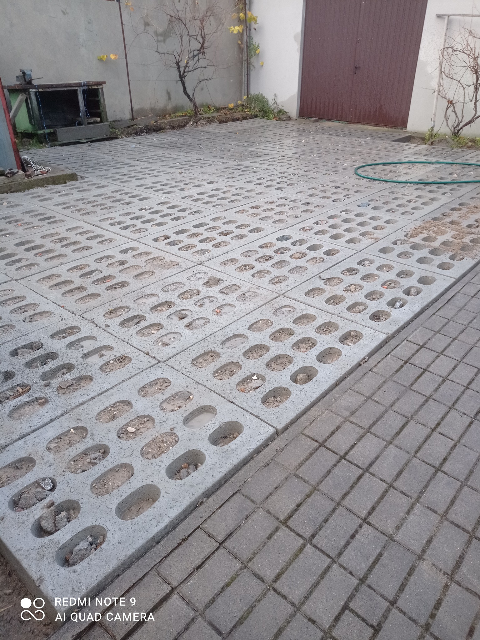 Ułożona szara ażurowa kostka betonowa z wypełnieniem w postaci ziemi i drobnych kamieni, widoczny fragment klasycznej kostki brukowej, zielony wąż ogrodowy i brama garażowa w tle.