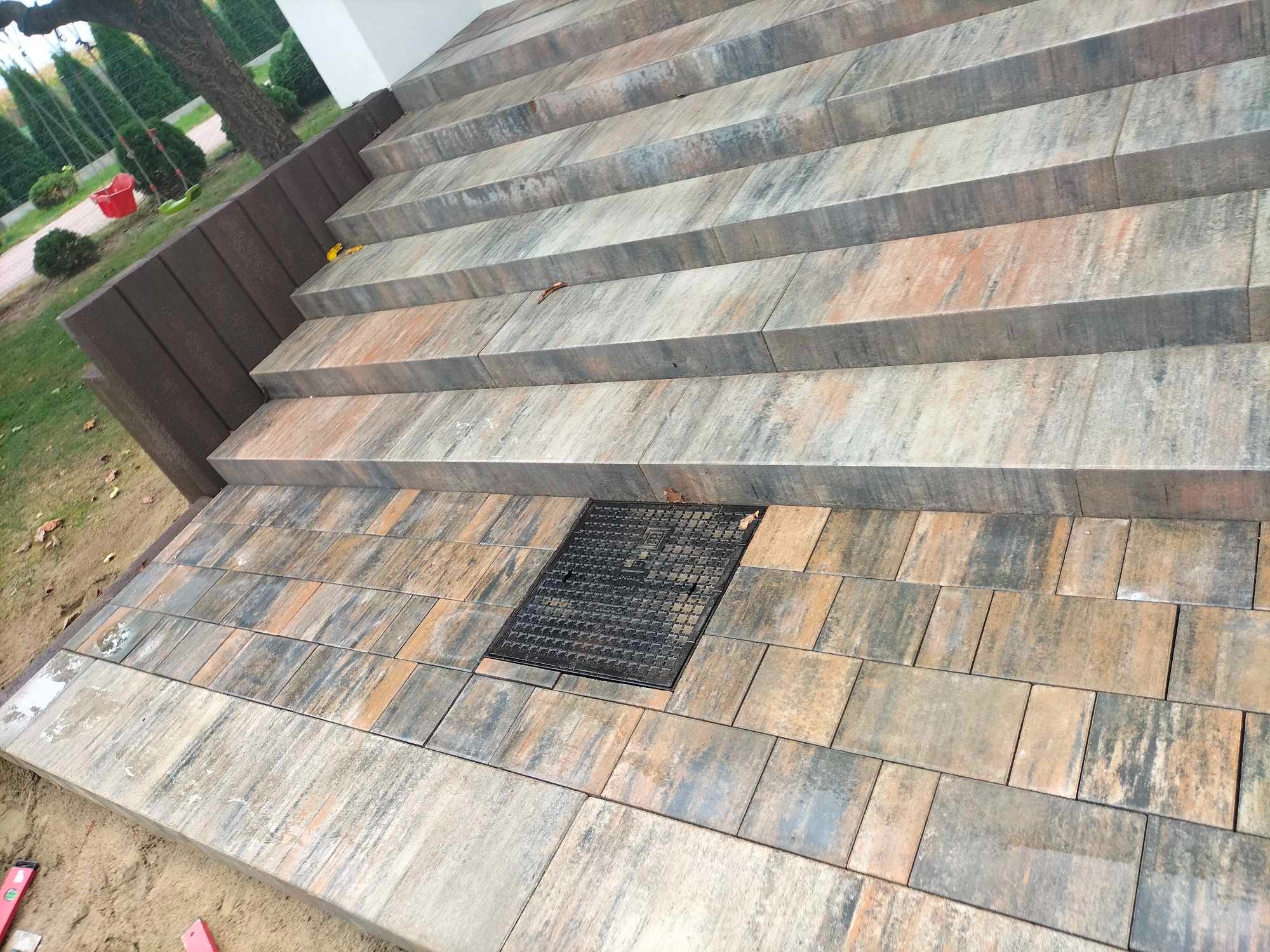 Schody i chodnik wyłożone wielobarwną kostką brukową z czarną studzienką kanalizacyjną pośrodku, widok z góry.