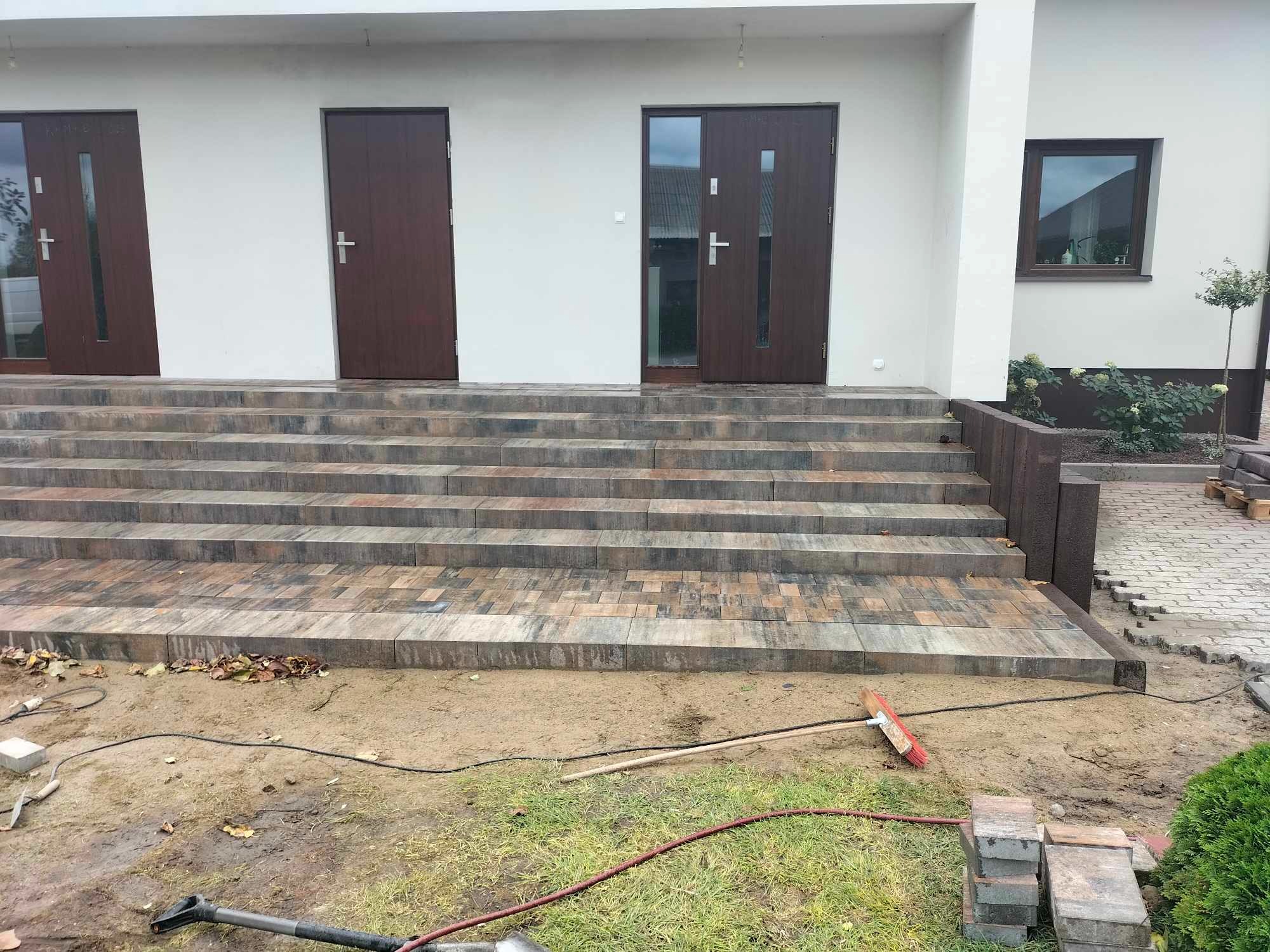 Schody wejściowe do domu wyłożone kostką brukową w odcieniach szarości i brązu, z widocznymi narzędziami brukarskimi i niezakończonymi pracami ziemnymi wokół.