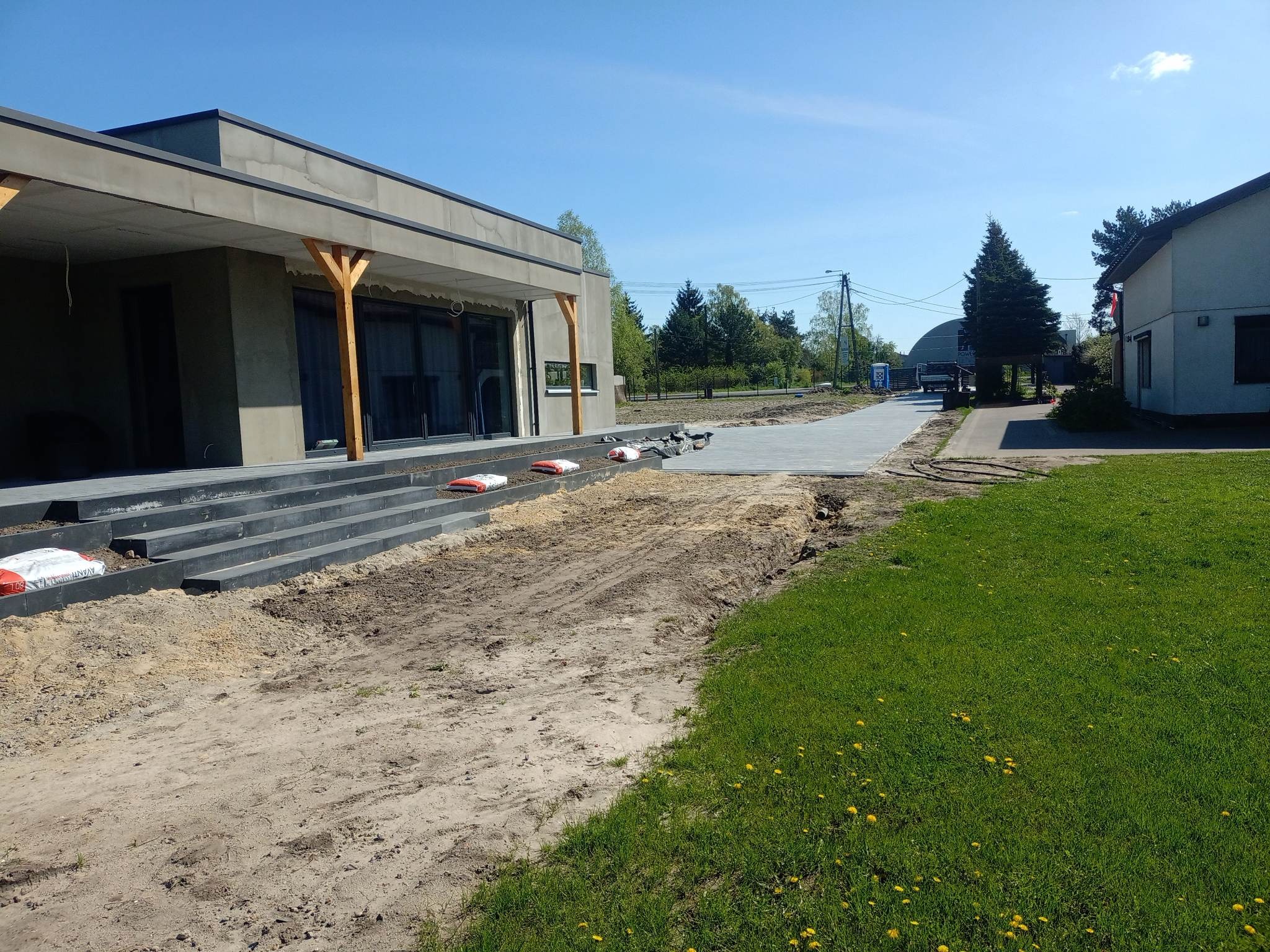 Ułożona, szara kostka brukowa tworząca ścieżkę i schody prowadzące do nowoczesnego domu z drewnianymi elementami, worki z materiałem budowlanym leżące na schodach, przygotowany teren pod dalsze...