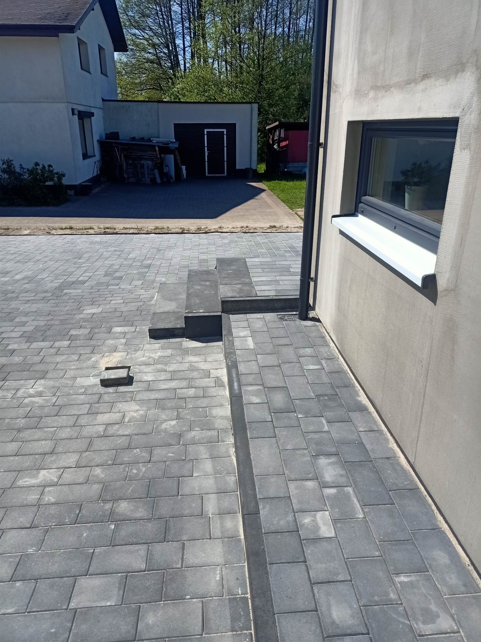 Ułożona szara kostka brukowa na podjeździe z widocznymi schodami i odwodnieniem liniowym przy ścianie budynku.