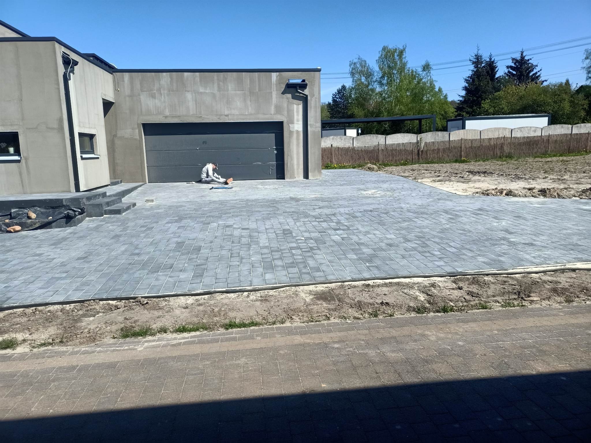 Ułożona szara kostka brukowa przed nowoczesnym domem z garażem; pracownik na kolanach, układający ostatnie elementy nawierzchni, widoczne narzędzia i piasek do fugowania.