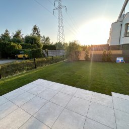 Usługi Ogrodniczo- Brukarskie Gardenin - Wykończony ogród z jasną, kwadratową nawierzchnią z płytek, równo przystrzyżonym trawnikiem, młodymi drzewkami i słupem wysokiego napięcia w tle, oświetlony zachodzącym słońcem.