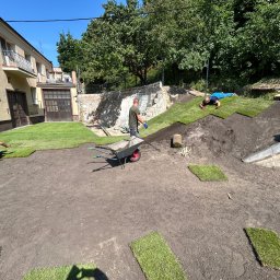 Usługi Ogrodniczo- Brukarskie Gardenin - Układanie darni w rolkach na przygotowanym terenie, widoczni trzej pracownicy, taczka z narzędziami i częściowo ułożona powierzchnia trawnika w ogrodzie przy budynku mieszkalnym.