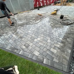 Usługi Ogrodniczo- Brukarskie Gardenin - Układanie kostki brukowej na posesji, widoczne świeżo ułożone szare kostki, obrzeże z kamyków, fragment trawnika, pracownik spłukujący powierzchnię wodą, ubijarka wibracyjna, oraz narzędzia pracy.
