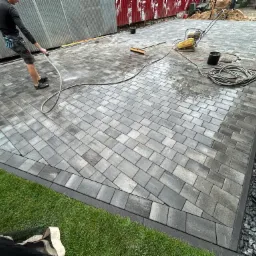 Układanie kostki brukowej na posesji, widoczne świeżo ułożone szare kostki, obrzeże z kamyków, fragment trawnika, pracownik spłukujący powierzchnię wodą, ubijarka wibracyjna, oraz narzędzia pracy.