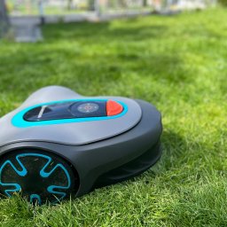 Usługi Ogrodniczo- Brukarskie Gardenin - Szaro-turkusowy robot koszący Gardena leży na gęstej, zielonej trawie w słoneczny dzień.