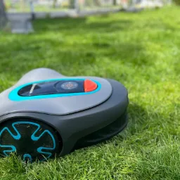 Szaro-turkusowy robot koszący Gardena leży na gęstej, zielonej trawie w słoneczny dzień.