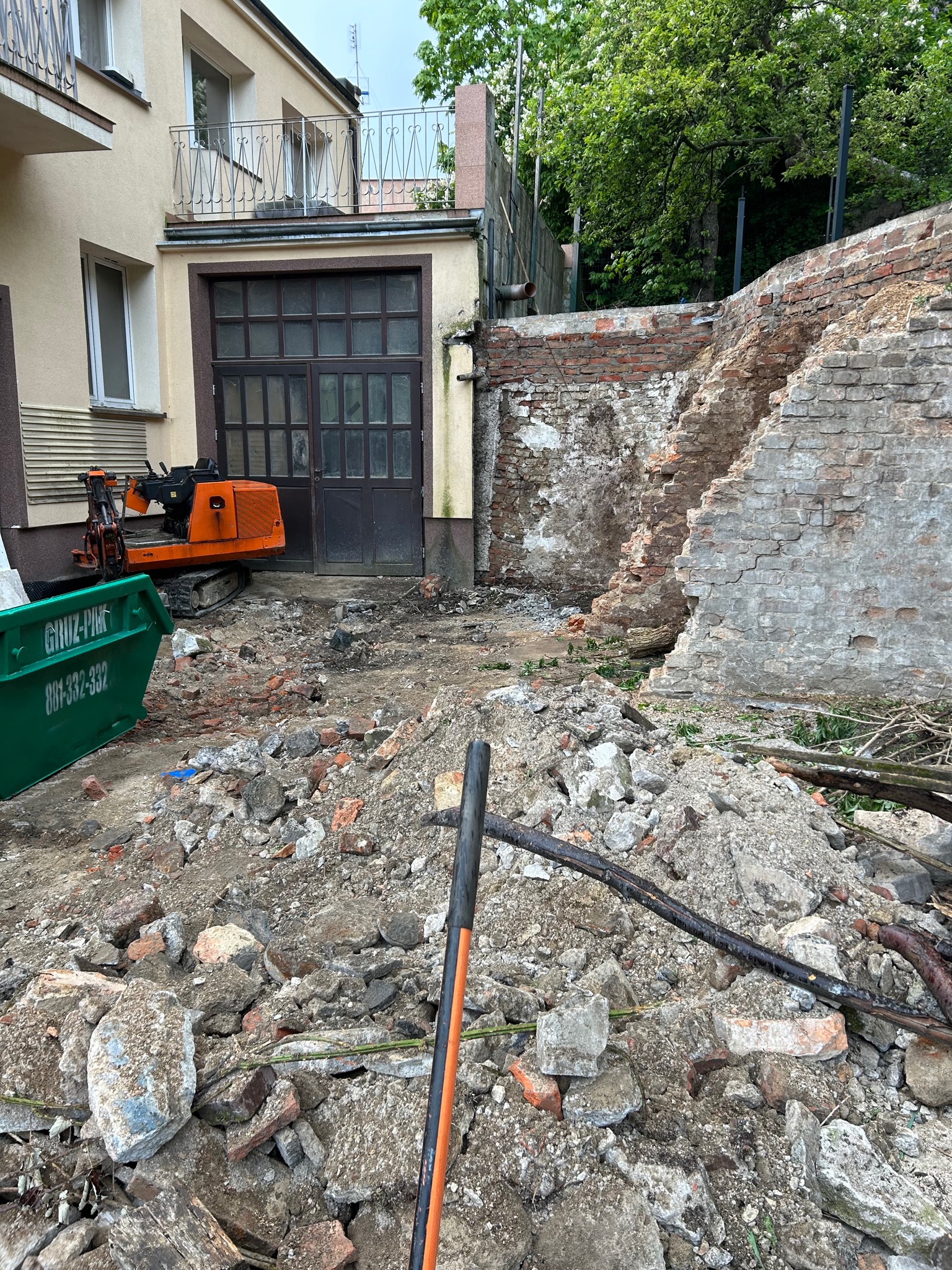 Pomarańczowa mini-koparka obok zielonego kontenera na gruz w Szczecinie, na tle częściowo zburzonej ceglanej ściany i budynku z garażem, widok na plac budowy.