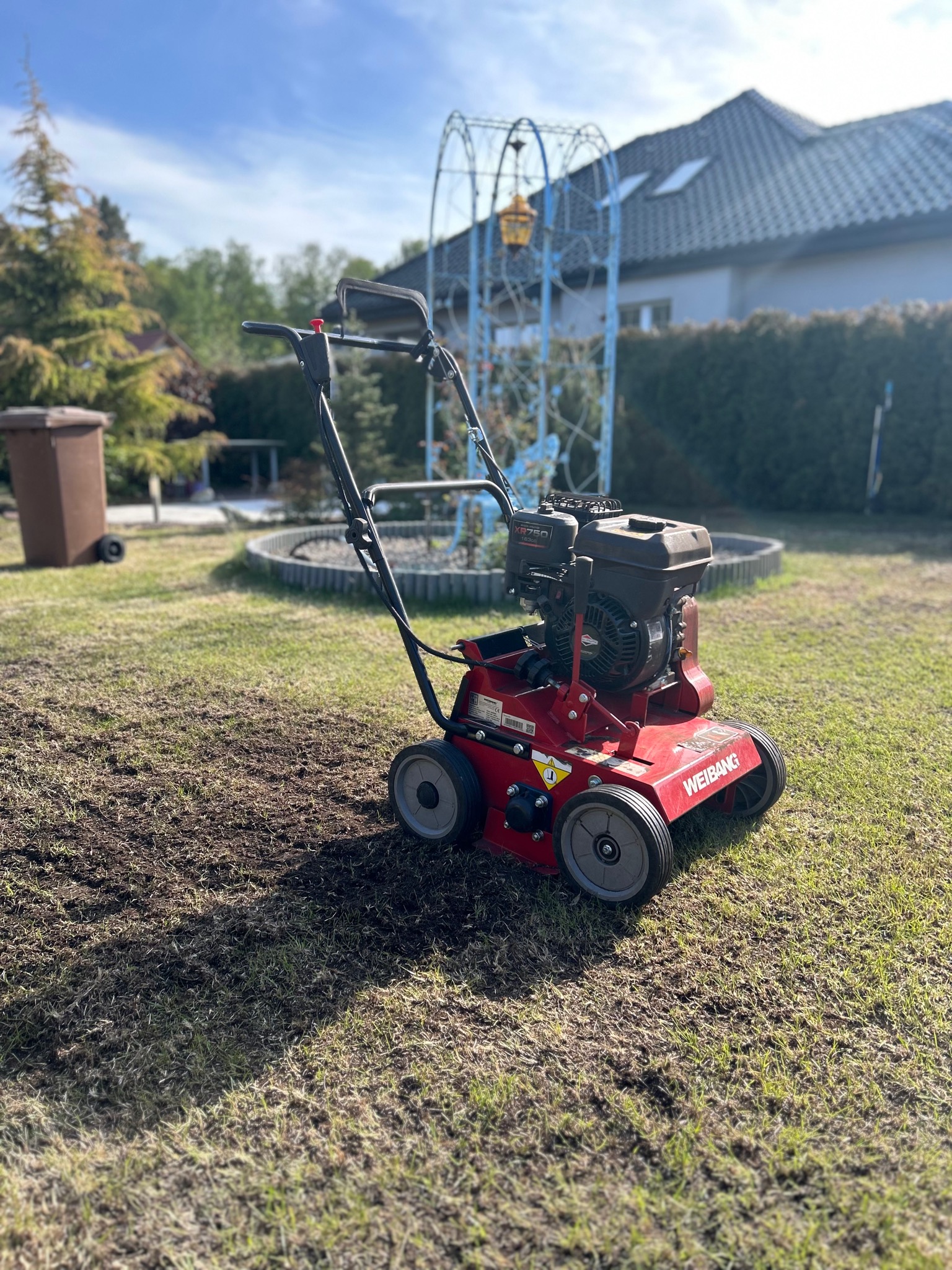 Czerwony aerator spalinowy Weibang na trawniku z widocznymi śladami aeracji, w tle dom z szarą dachówką i dekoracyjna konstrukcja ogrodowa.