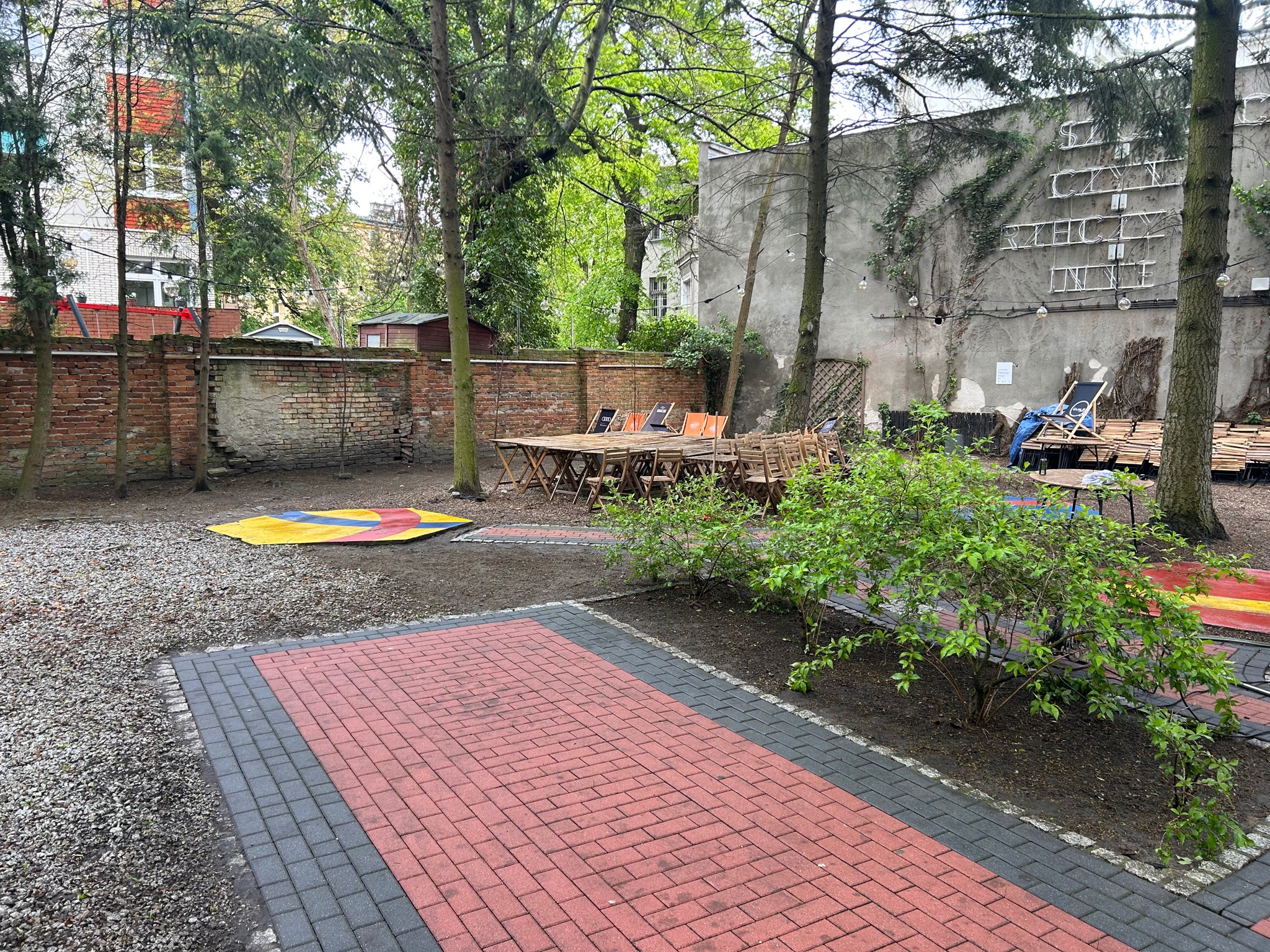 Ułożona czerwono-szara kostka brukowa w ogrodzie z drzewami, stolikami i neonem na ścianie w tle.